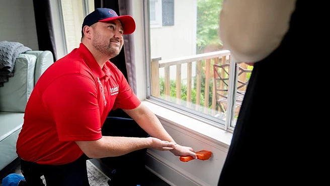 A Rainbow Restoration service professional bending down and using a moisture meter on a wall below a large window, showing the reading to a customer.