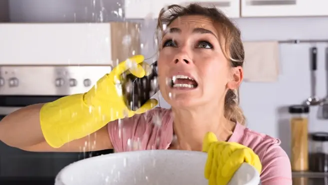 A woman holds a bucket to catch water coming from the ceiling while on the phone calling a water damage restoration company for emergency service.