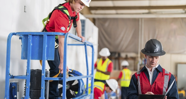 Workers in safety gear on a jobsite. One man writes on a clipboard. Another preps a hydraulic lift.