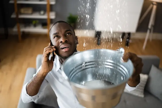 Galveston homeowner calls for water damage restoration help while holding a bucket collecting water coming from the ceiling.