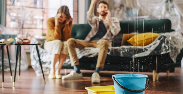 A man and woman sit on a sofa to call a water damage restoration company in Oskaloosa, IA, as water falls from the ceiling and collects in a container on the floor.