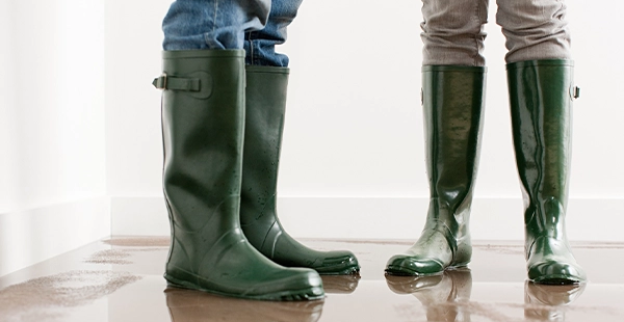 A couple in Deschutes River Woods, OR, wears rubber boots and stands in a flooded living room surrounded by water-damaged carpet.