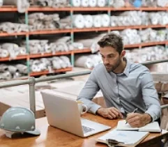A manager looking at a laptop at a table in a warehouse.