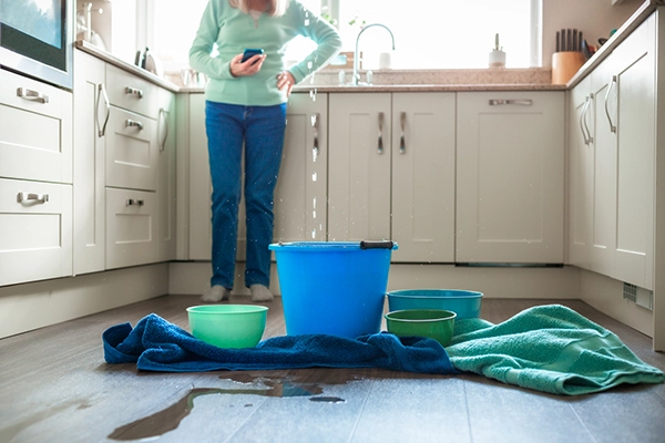 Woman in Five Forks, SC, stands in the kitchen holding a phone to call a restoration company during a water damage emergency as water falls from the ceiling.
