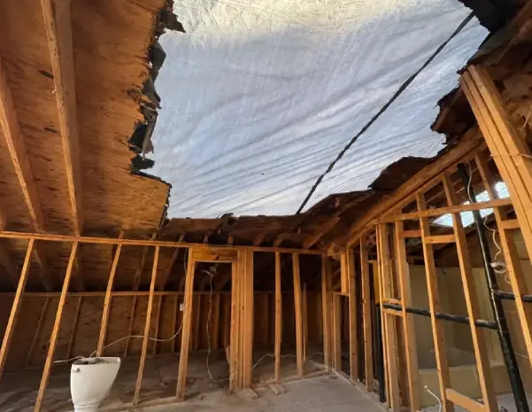 A storm-damaged roof is temporarily covered with a protective tarp above an exposed residential interior.