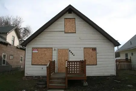 A white two-story house with boarded-up windows and doors, showing plywood coverings over all openings.