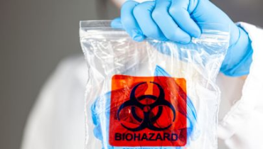 A person wearing blue nitrile gloves and a white protective suit holds a clear plastic biohazard specimen bag. The bag features a prominent orange biohazard symbol and contains medical or contaminated materials, indicating safe handling during a biohazard cleanup process.