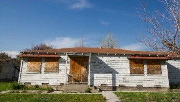A single-story white house with boarded-up windows and front door, indicating damage or vacancy.