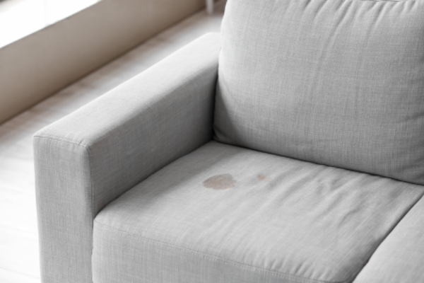 A light gray upholstered sofa with a noticeable dark stain on the seat cushion, showing the kind of fabric damage that requires professional upholstery cleaning.