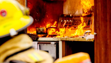 A firefighter in the foreground with a burning kitchen in the background.