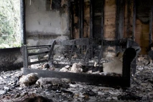 A severely fire-damaged living room with charred walls, a burned sofa frame, and debris scattered across the floor. The structure shows heavy smoke staining and destruction, highlighting the aftermath of a house fire.